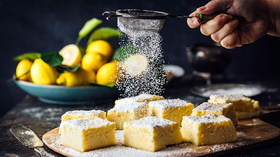 Seitenansicht einer weiblichen Hand, die Puderzucker über Zitronenkuchenquadrate streut