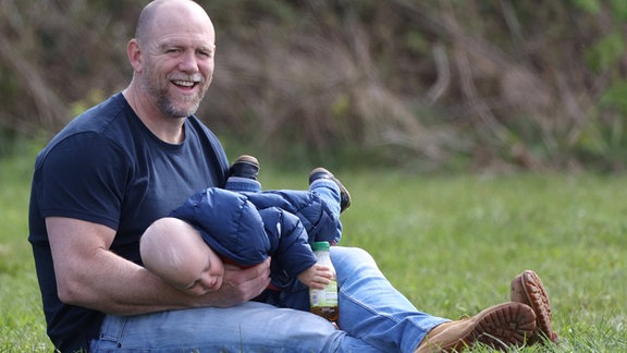 Mike Tindall sitzt mit einem Kind auf einer Wiese.